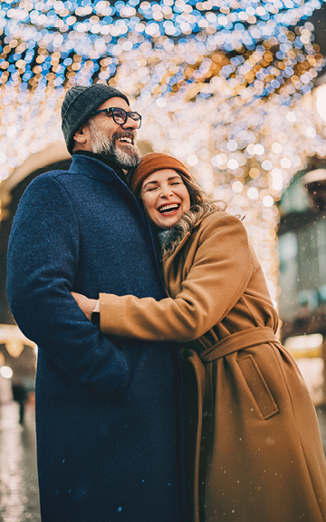 Ein älteres Paar steht vor einem glitzernden Hintergrund und umarmt sich. Beide tragen Winterkleidung und lächeln.