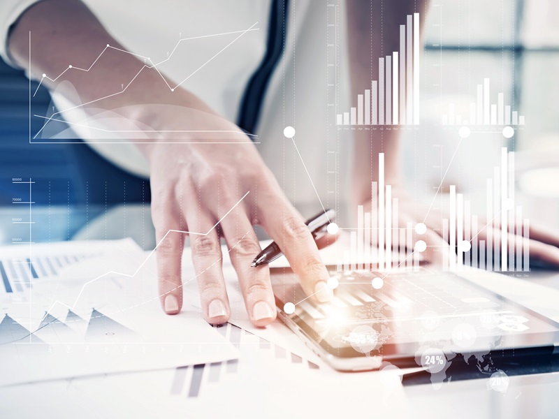 Eine Hand tippt auf einem Tablet. Im Vordergrund sieht man leicht transparent verschiedene Grafiken und Linien.