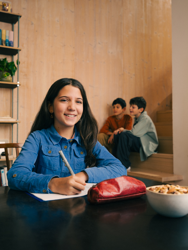 Ein Mädchen sitzt an einem Tisch bei den Hausaufgaben. Sie hält einen Stift in der Hand und lächelt in die Kamera.