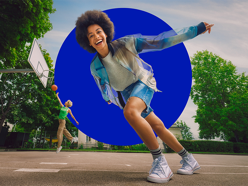 Zwei junge Frauen spielen Basketball. Eine der Frauen wirft im Springen einen Korb, die andere läuft an der Kamera vorbei und lacht.