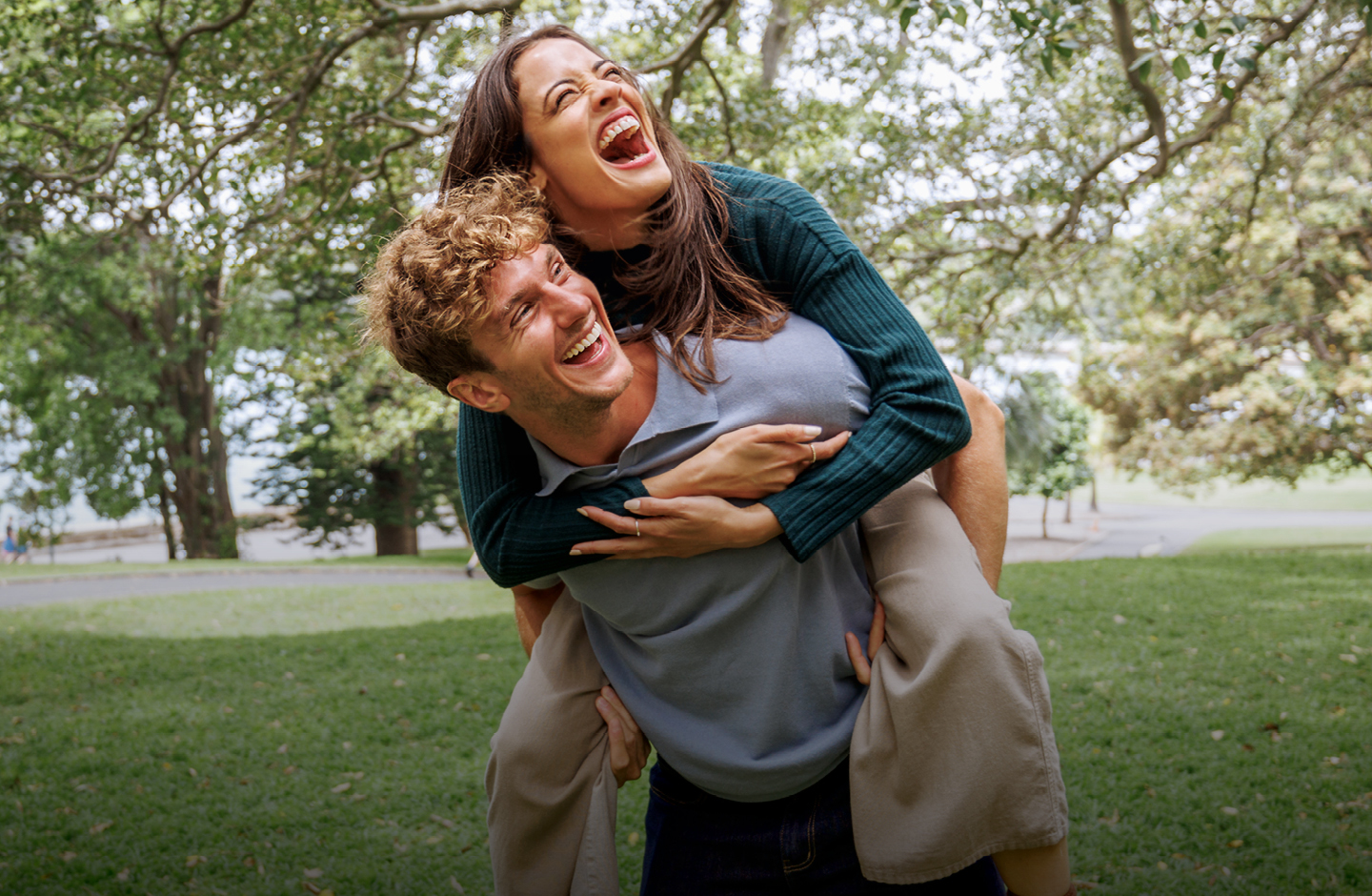 Ein Mann und eine Frau sind in einem Park an einem See. Er hat sie huckepack. Sie lachen beide ausgelassen.