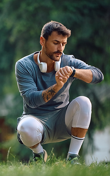 Ein Mann in Sportkleidung und mit Kopfhörern um den Hals ist in der Hocke und bedient seine Smart-Watch.