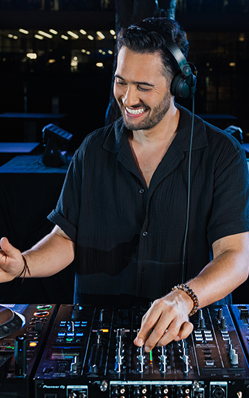 Ein DJ in einem schwarzen Hemd steht for seinen Turntables und legt Musik auf. Er hat Kopfhörer auf und lacht.