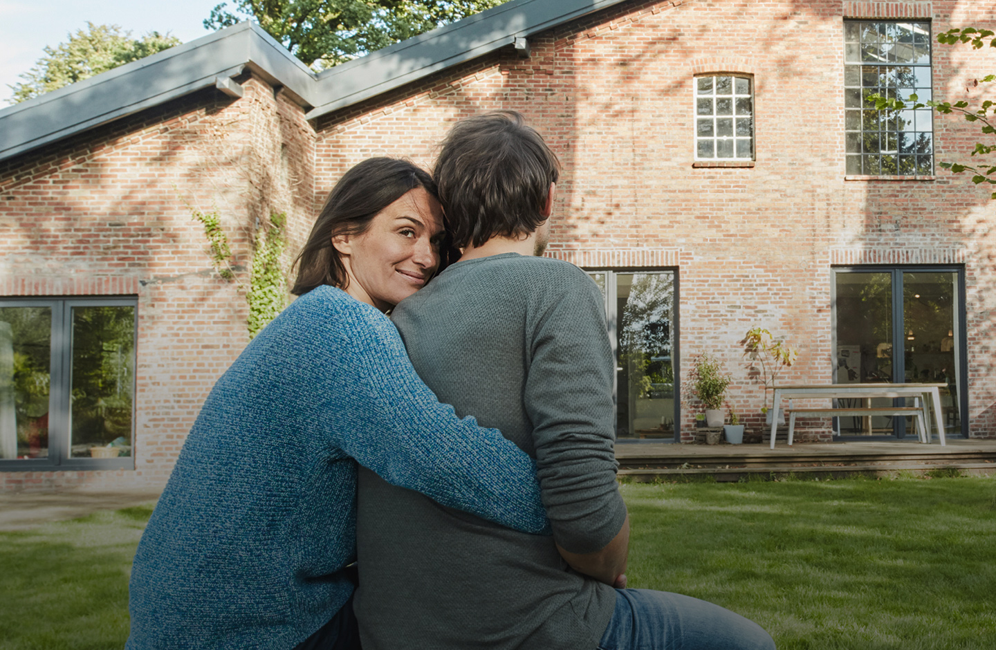 Ein Mann und eine Frau sitzen mit dem Rücken zur Kamera vor einem Haus. Die Frau umarmt den Mann von hinten. Sie blickt über die Schulter in die Kamera.