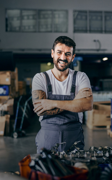 Ein Mann in Latzhose steht in einer Werkstatt und verschränkt lachend die Arme.